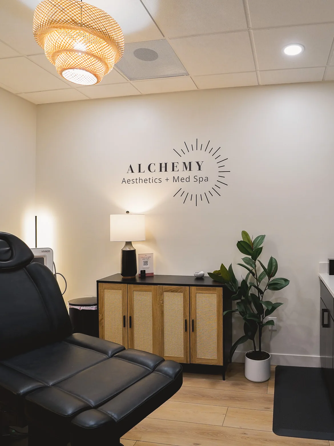 A treatment room at Alchemy Aesthetics and Med Spa, with the clinic name on the wall, a treatment chair and a warm lamp.